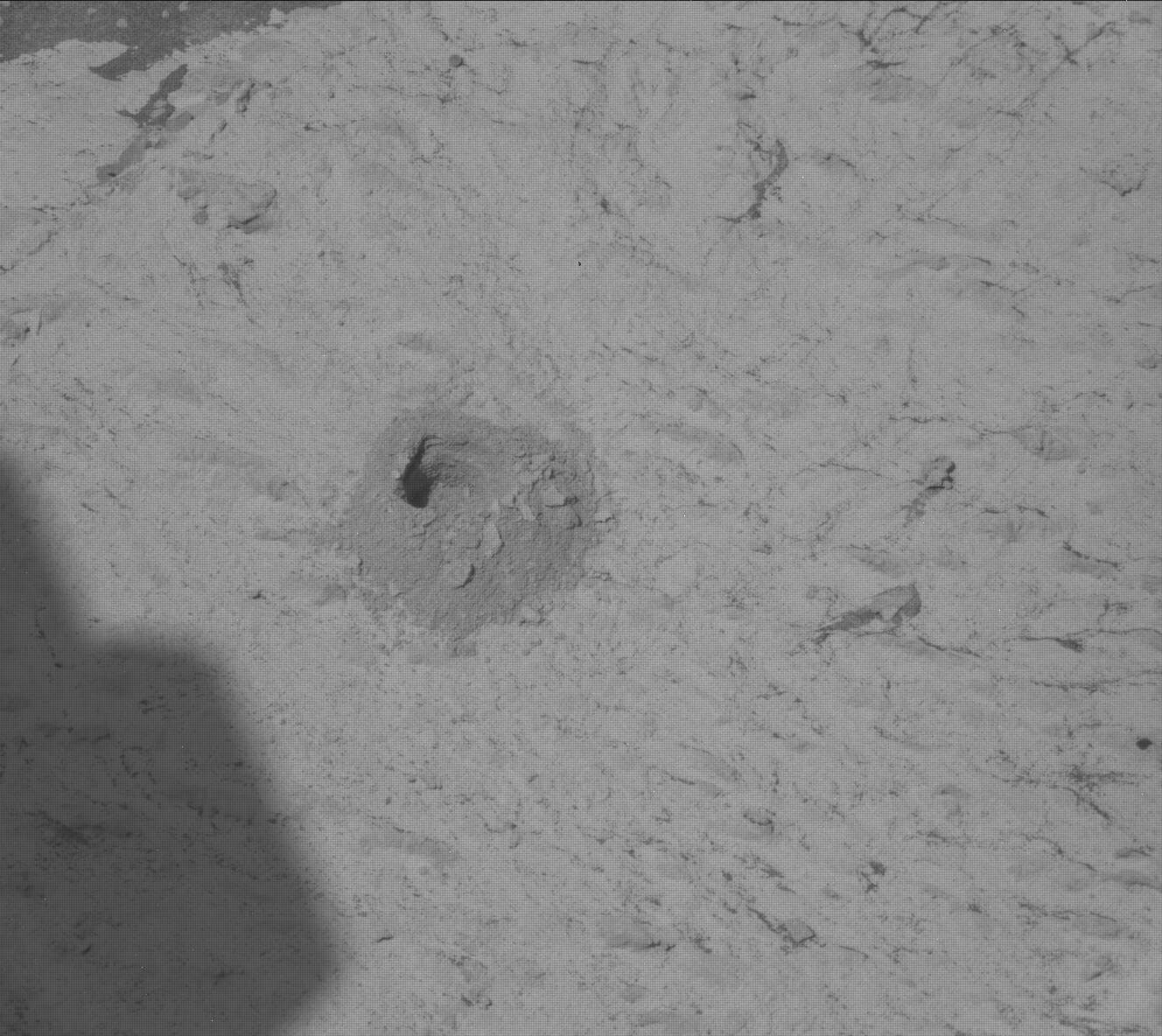 This black and white image shows a quarter-size drill hole on the rocky, sandy surface of Mars. Curiosity's shadow is also present in the bottom left corner of the image. 