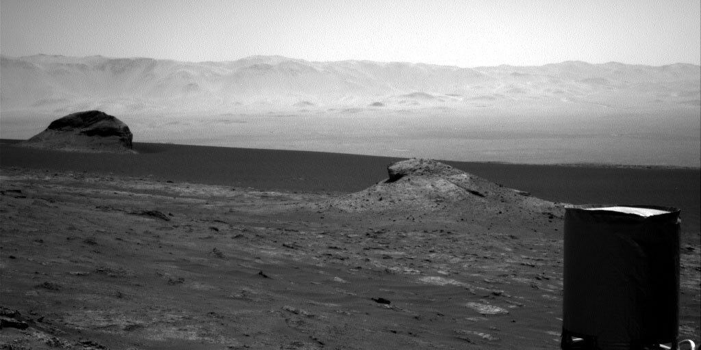 This is a black and white image of the rocky surface on Mars. In the center of the image there is a small dune of sand and rock.There is also another much darker rock to the left. In the background the smoothness of the sand and hills is displayed. 