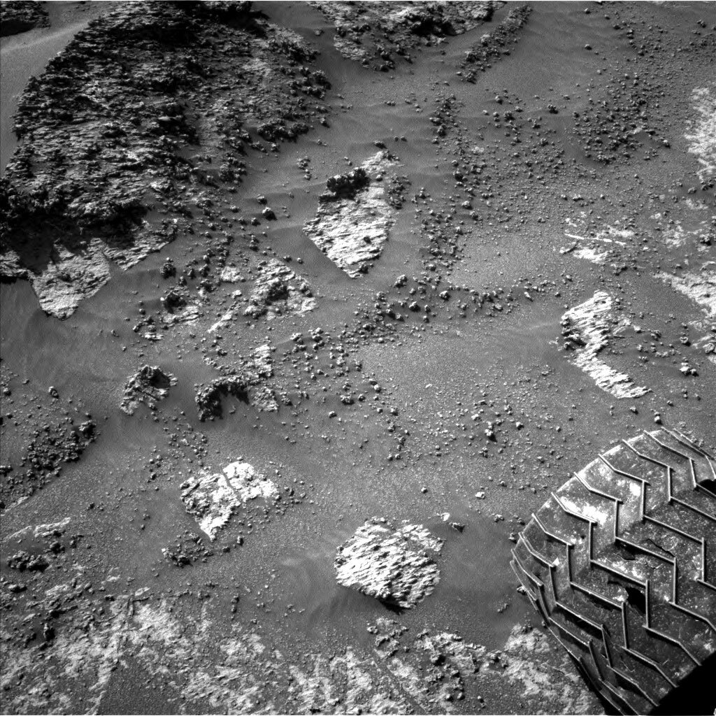 This is a black and white image of the rocky surface of Mars. There is a very rough, textured large rock in the top right corner of the image. The surface surrounding the rock is smooth, sandy and dusty with small rocks scattered all over. Some parts of the rock are peering through the sand in the center and the bottom of the image. Curiosity’s left tire can be seen in the bottom right crooner of the image. 
