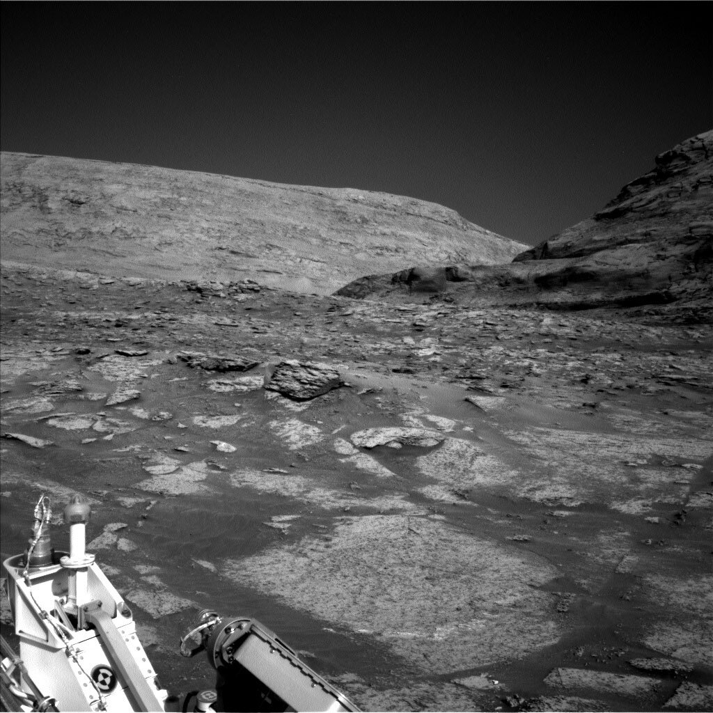 This is a black and white image of the rocky, sandy surface of Mars. In the front view rocks are embedded in sand beneath the surface. Two large hills and a clear sky are in the horizon of the image. 