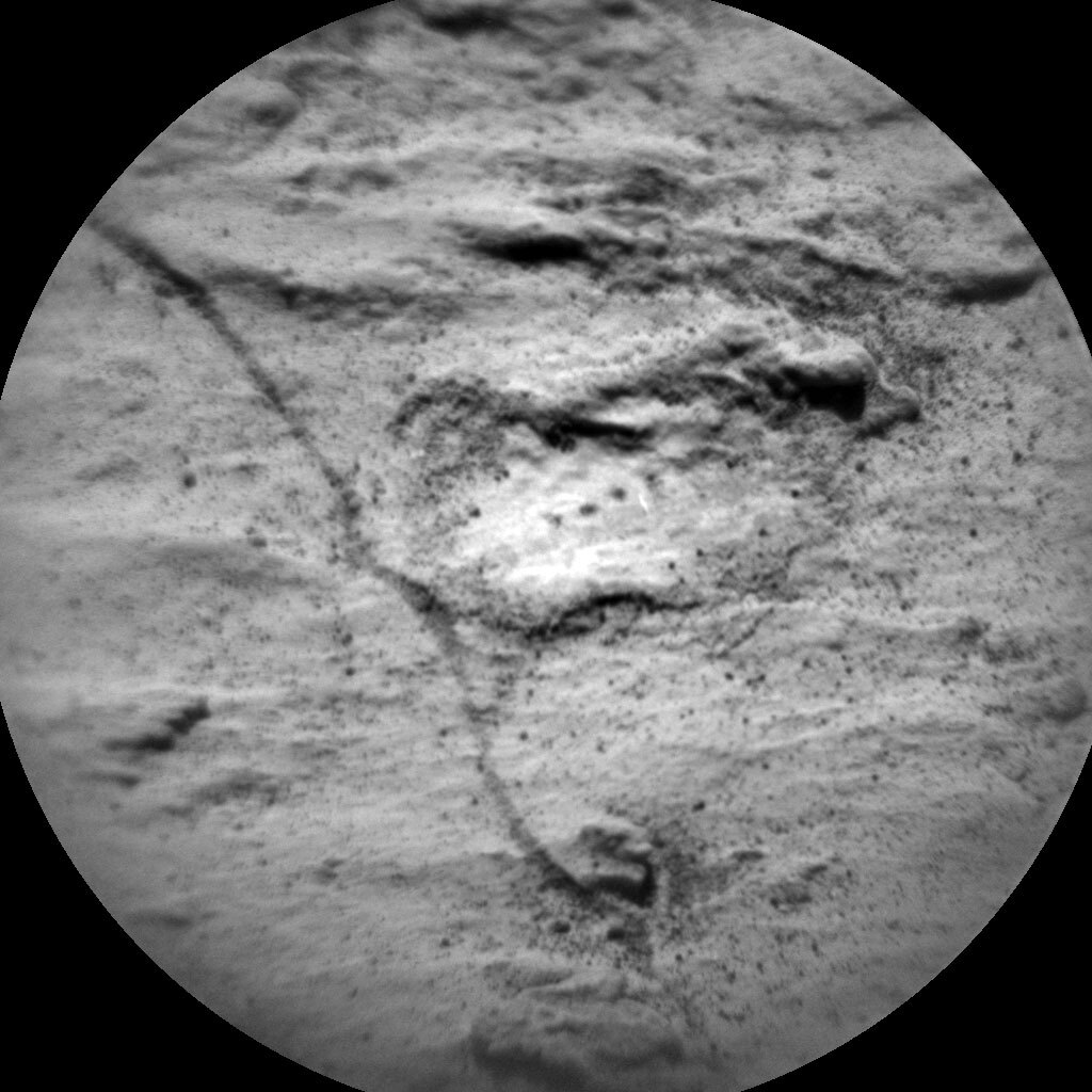 This is a black and white circular lens image of the rough and rocky surface of Mars. The surrounding are to the roughness is smooth, flat and has little rock particles scattered on it. 