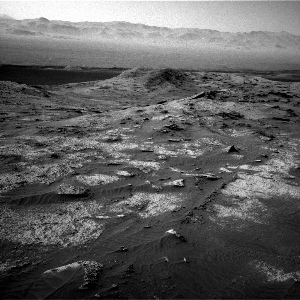 This is a black and white image of the rocky, sandy surface on Mars. In the front of the image rocks are seen embedded into the ground and are slightly poking out in places. Larger hills can be seen in the horizon with a clear sky.  