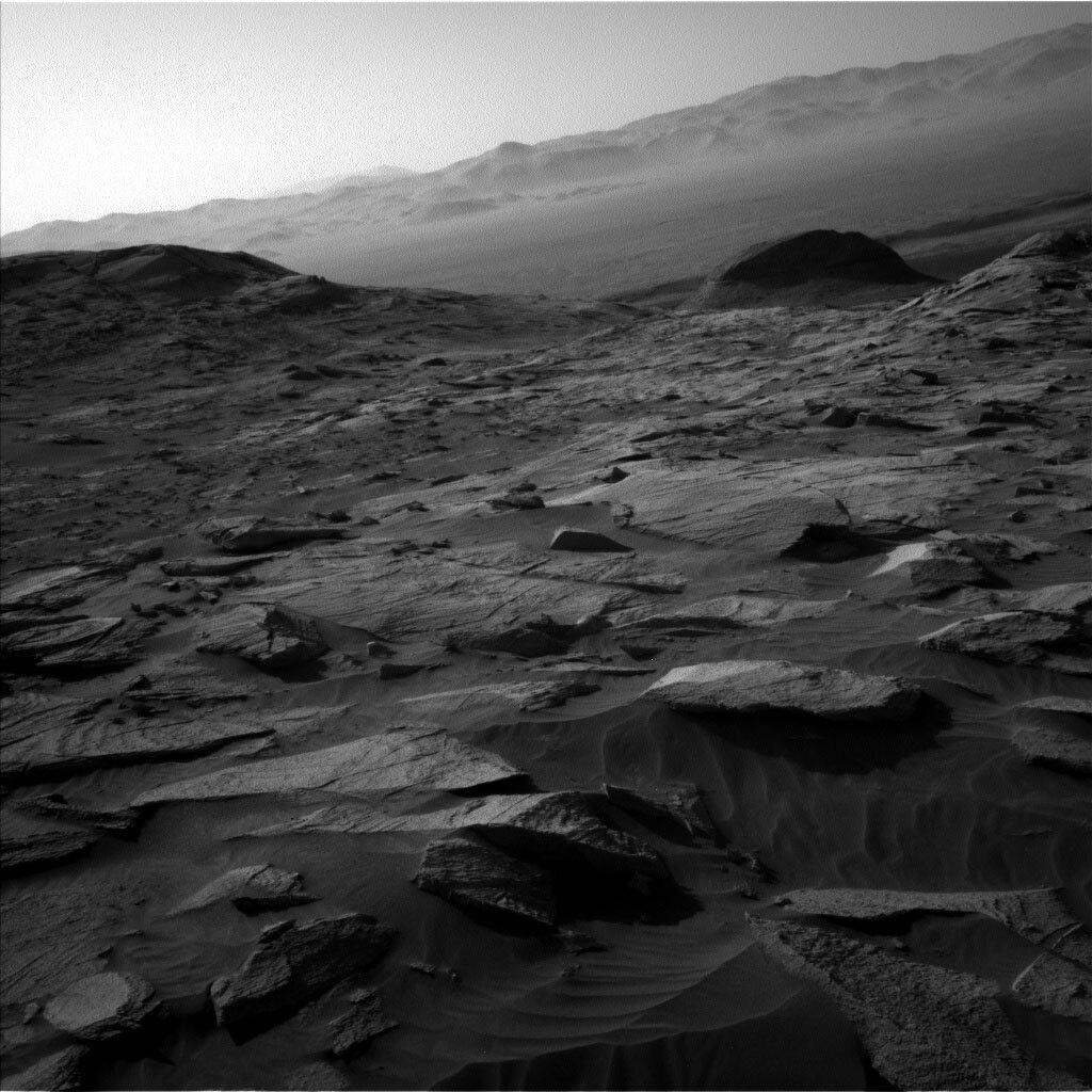 This is a black and white image of large boulders and hills in the horizon. The texture over the rocks is smooth sand.  