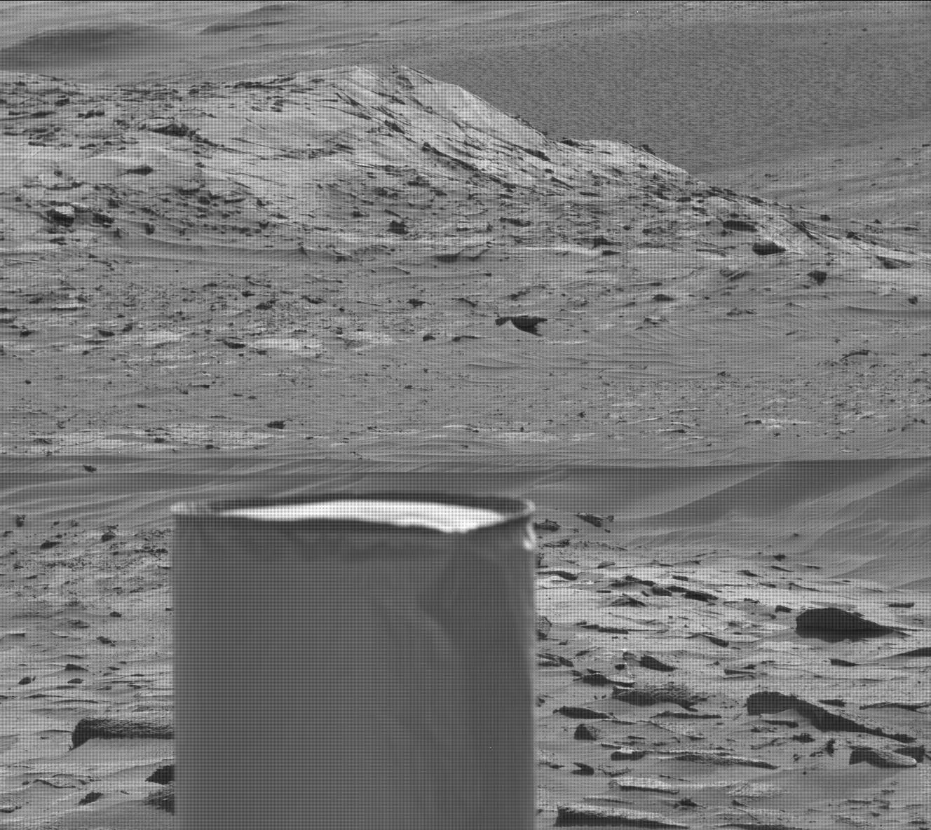 This is a black and white image of the smooth, sandy surface of Mars. There are two low hills in the background with lots of small scattered boulders. 