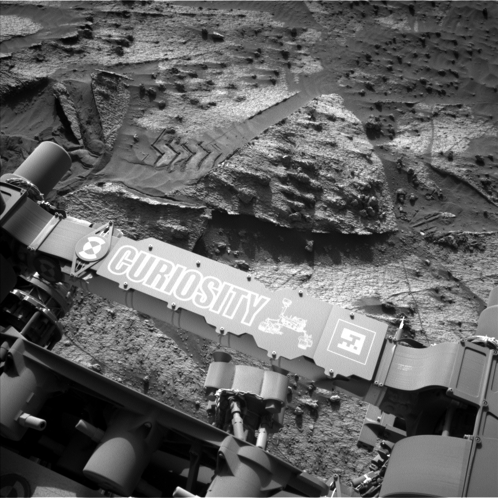 This is a black and white image of the Curiosity rover over the sandy, rocky surface of Mars. Curiosity's arm and tire marks can be seen imprinted in the smooth sand. 