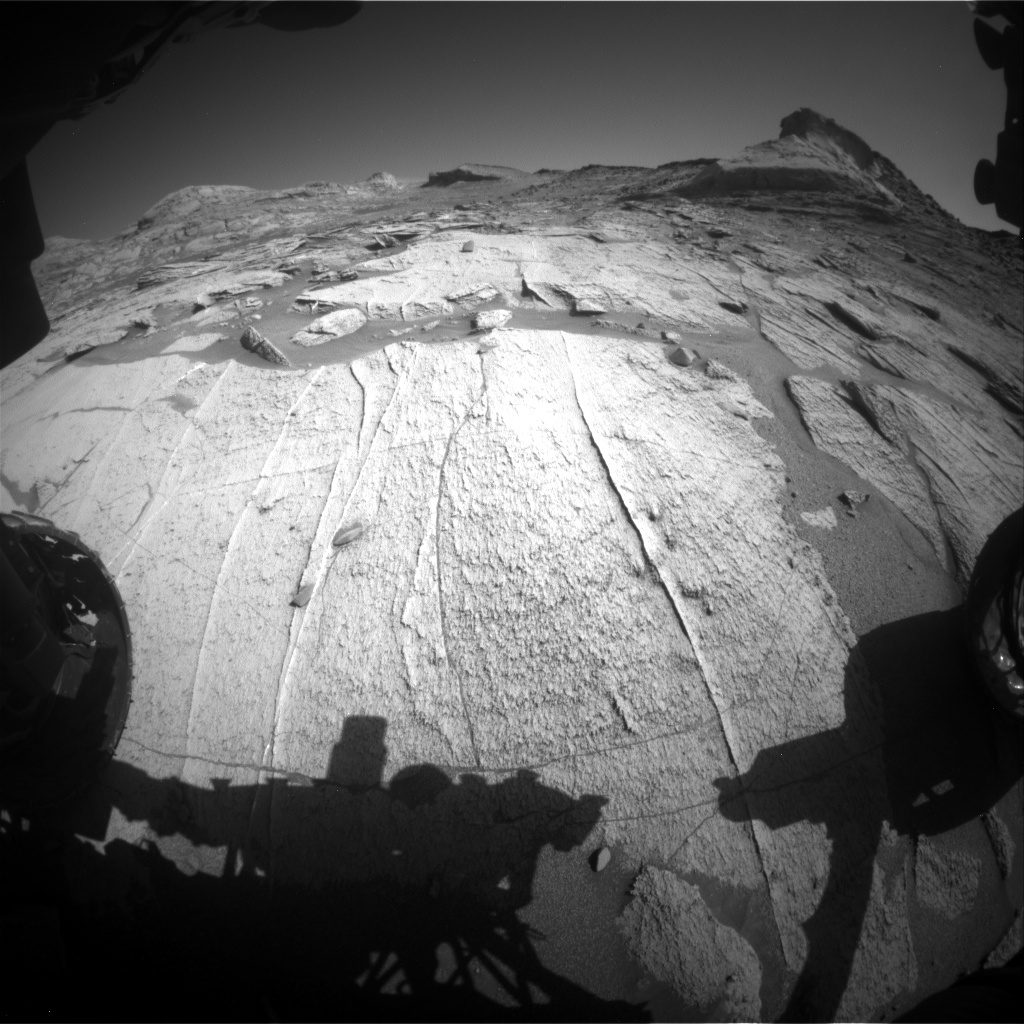 The Curiosity rover’s tires and shadow are in the forefront of the image. This is a black and white image showing the rocky, sandy surface of Mars. Low hills and a clear sky are in the horizon of the image. 