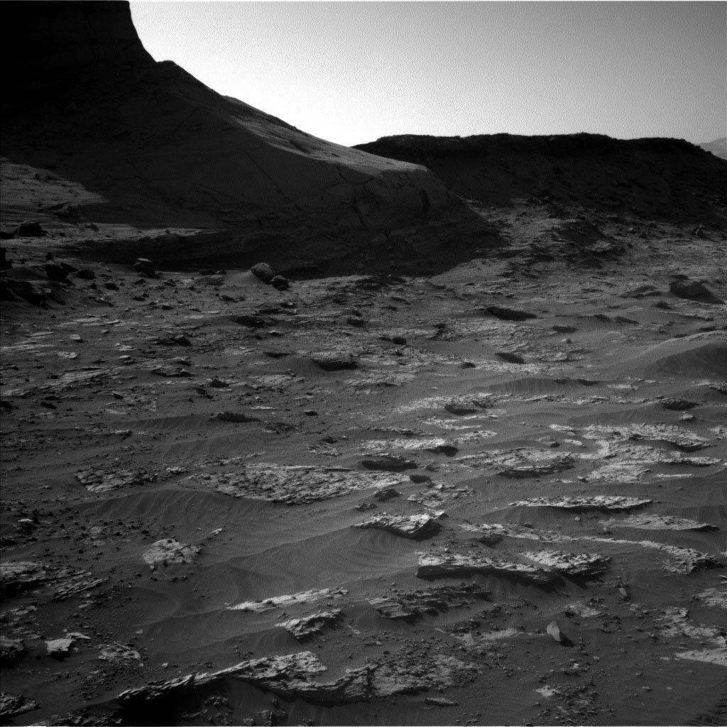 This is a black and white image of rocks embedded in sand on Martian ground. Low hills are present in the horizon of the image against a clear sky. In the forefront smooth sand and small scattered rock particles are present. 
