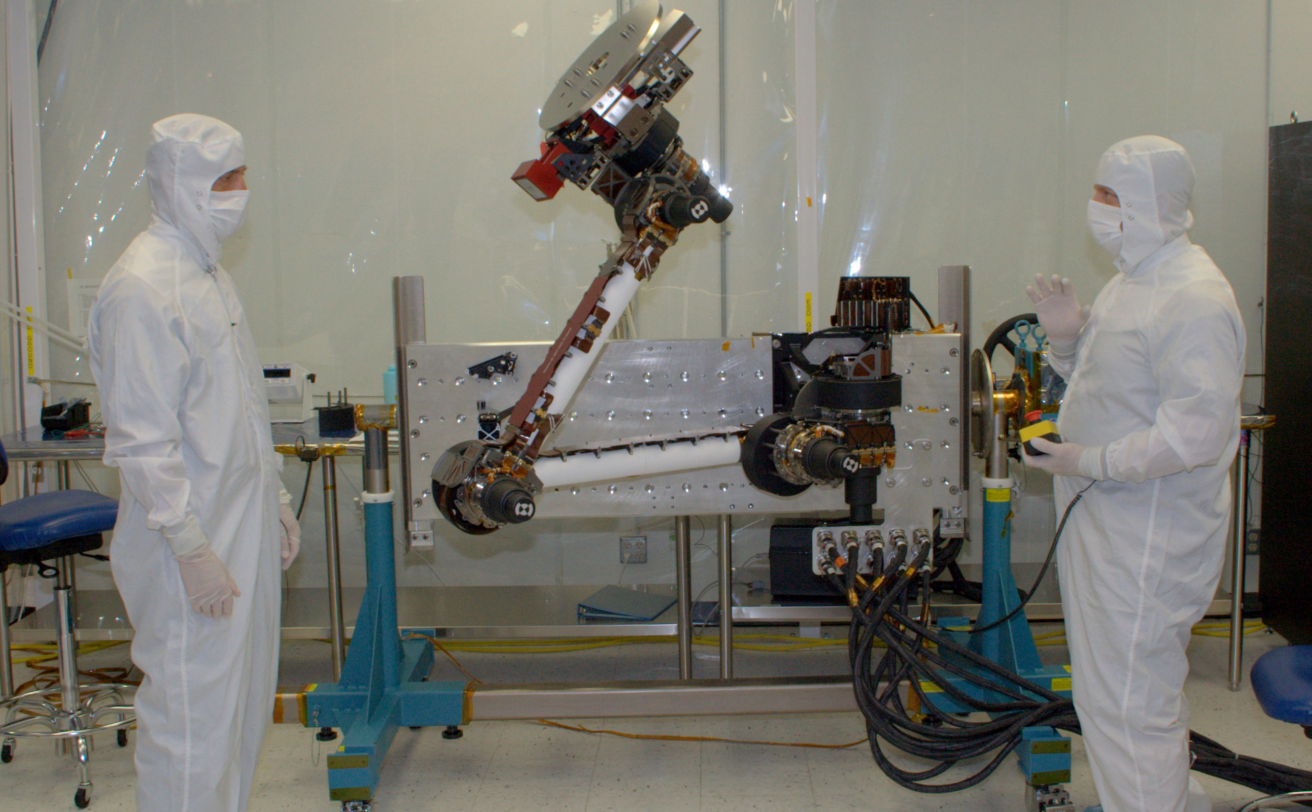 In this photo, two engineers from NASA Jet Propulsion Laboratory and Alliance Spacesystems are standing in a white room on either side of the Mars Science Laboratory rover's robotic arm. They are dressed in white 'bunny suits,' and are covered from head to toe wearing white face masks to protect the equipment. The arm is bent at the 'elbow' and raised high in the air about two feet higher than the engineer on the left. The engineer on the right holds a control box that maneuvers the arm for testing.